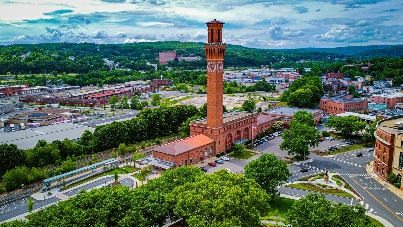 A tall brick clock tower stands amid a complex of brick buildings, surrounded by trees and roads, with a sprawling industrial and residential area in the background under a partly cloudy sky. Republican American clock tower in Waterbury, CT.