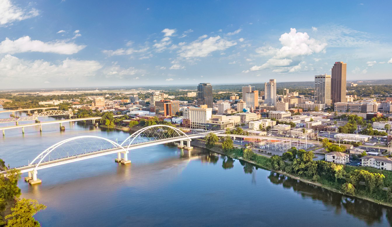 Aerial view of a city skyline with modern buildings, bridges crossing a wide river, and scattered clouds in a bright blue sky.