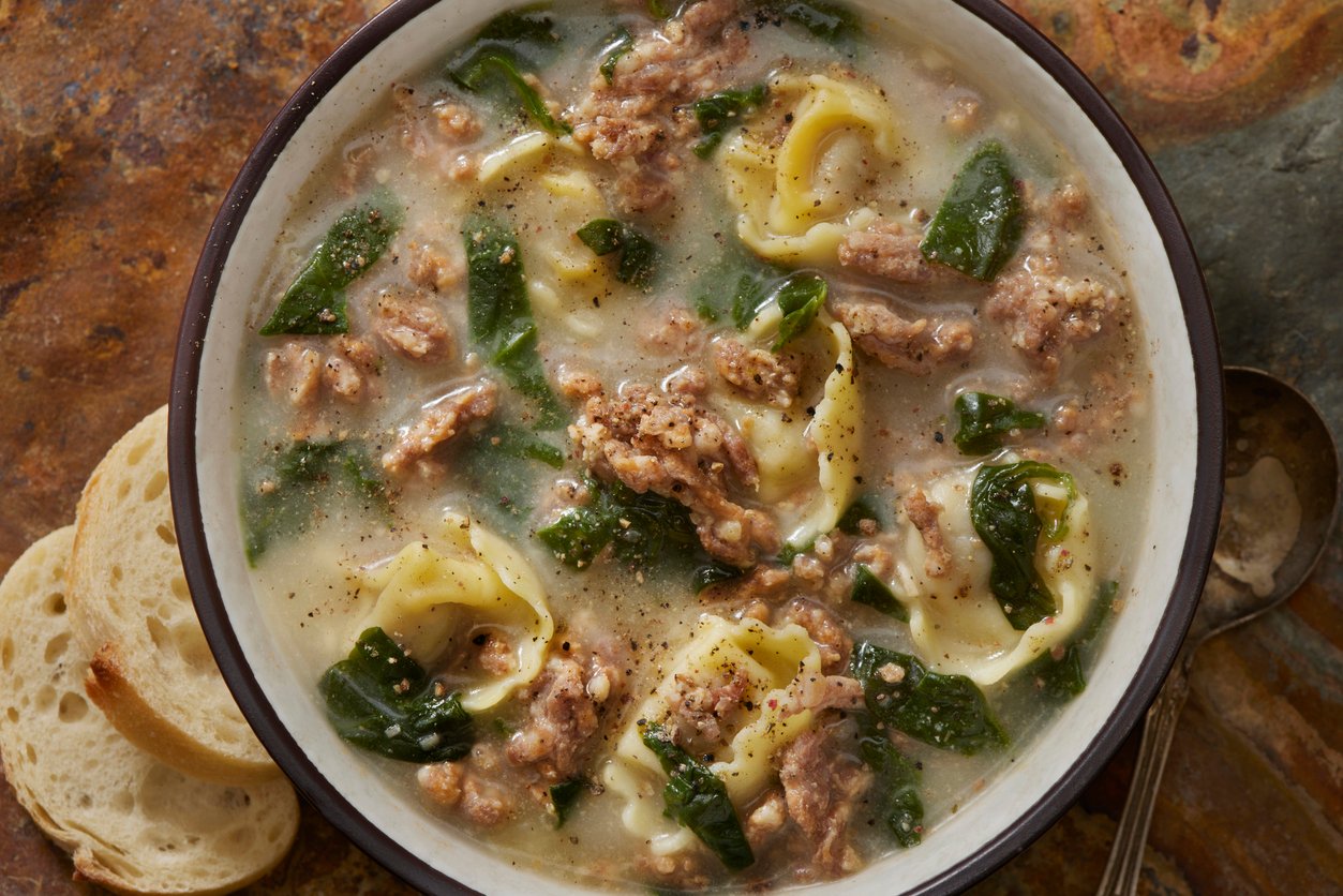 A bowl of creamy soup with tortellini, spinach, and ground sausage, topped with black pepper. Two slices of bread sit next to the bowl on a rustic surface, with a spoon partially visible.