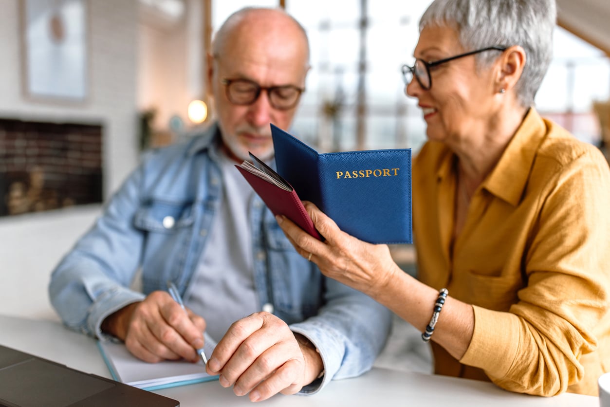 An older woman in glasses holds a blue passport and smiles at an older man, who is writing in a notebook. They sit together at a table in a bright, modern room.