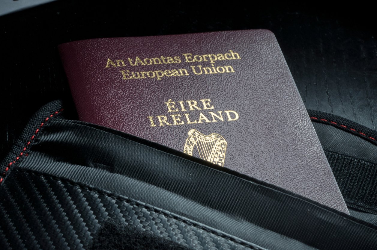 An Irish passport partially tucked into a black, textured bag with red stitching, showing both Irish and English text and an embossed harp emblem on the cover.
