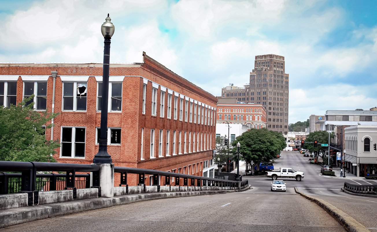 A view down a city street with red brick buildings, a tall tower in the background, trees along the sidewalks, and a few cars driving and parked in the area on a partly cloudy day.