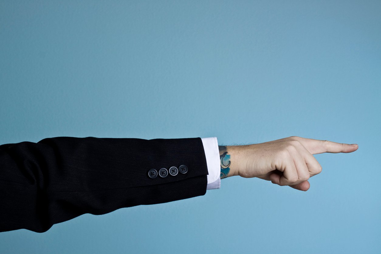 A person in a black suit points to the right with their index finger extended against a plain blue background. A blue-green tattoo is visible on their wrist.