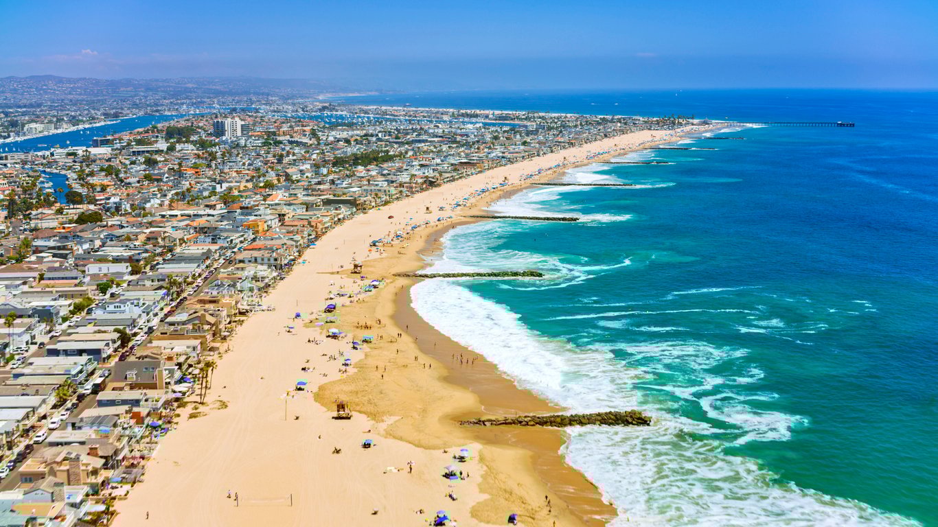Aerial view of a long sandy beach lined with houses and buildings, with people sunbathing and swimming. Waves roll onto the shore and a pier extends into the blue ocean under a clear sky.