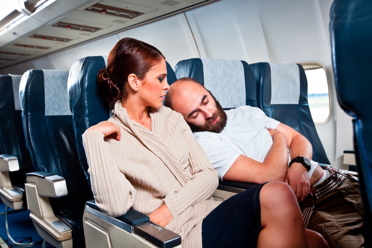 A woman sits in an airplane seat looking at a man next to her who is asleep and leaning on her shoulder. The plane seats are blue and grey, and the cabin appears calm and quiet.