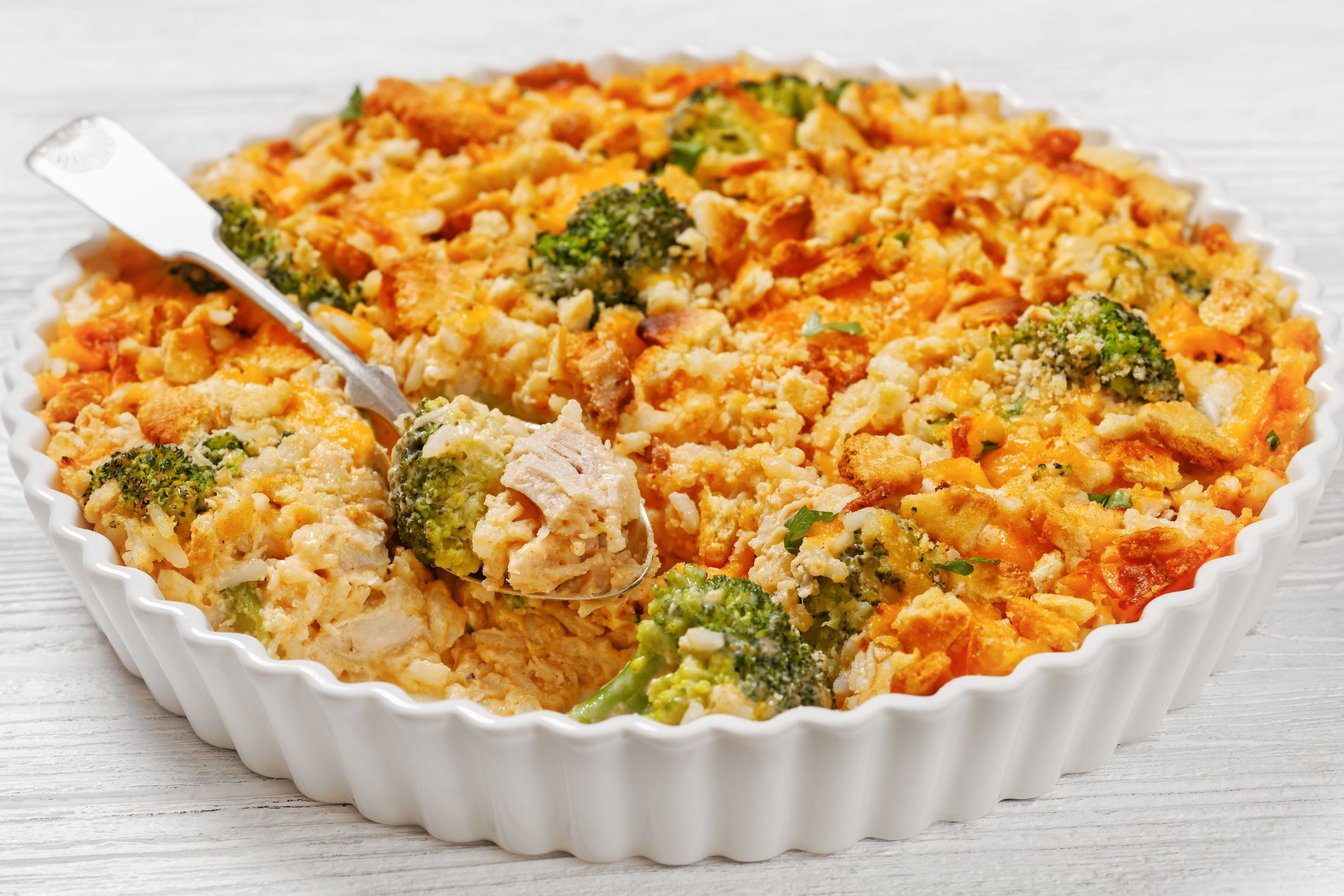chicken rice broccoli casserole topped with cheddar cheese and crumbled cracker in baking dish on white wooden table, landscape view from above