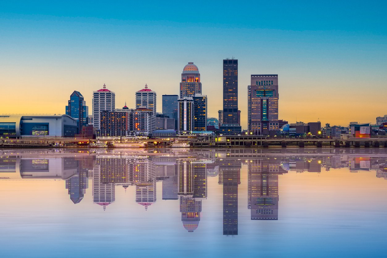 Louisville city skyline at sunset, with tall buildings and structures reflected clearly in a calm river, under a blue and orange sky.