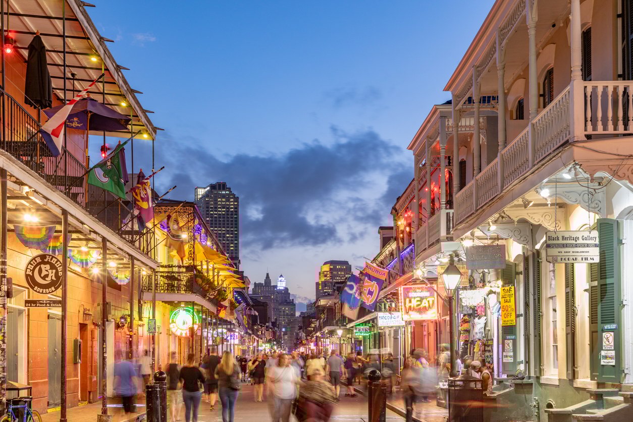 A lively street scene in the French Quarter of New Orleans at dusk, with colorful lights, flags, many people walking, and historic buildings lining both sides of the bustling street.