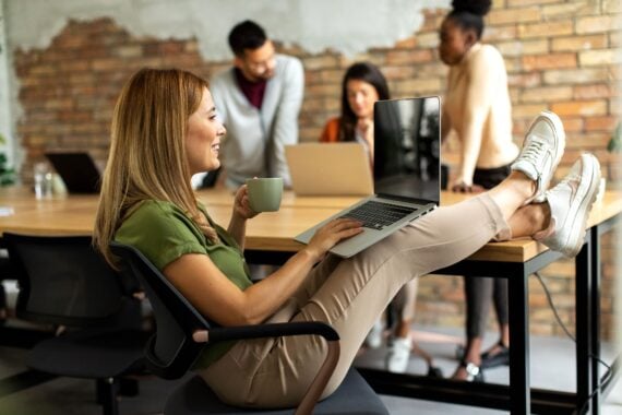 A woman sits at a desk with her feet up, holding a mug and using a laptop. In the background, three colleagues stand together, talking and working on laptops in a modern office with exposed brick walls.