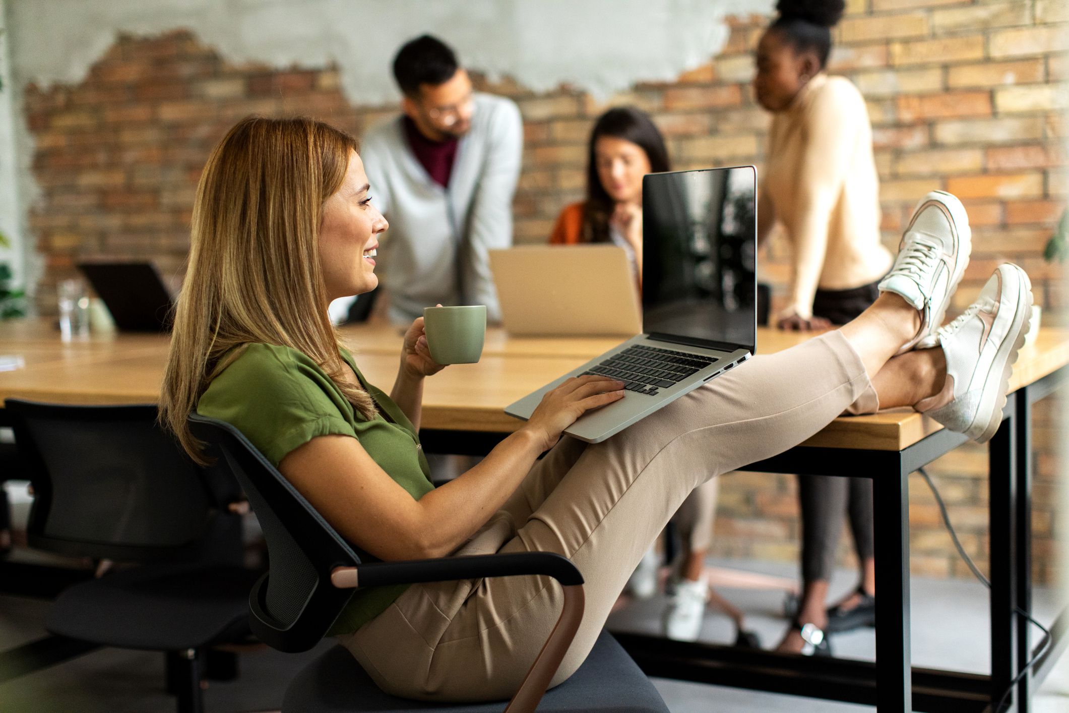 A woman sits at a desk with her feet up, holding a mug and using a laptop. In the background, three colleagues stand together, talking and working on laptops in a modern office with exposed brick walls.