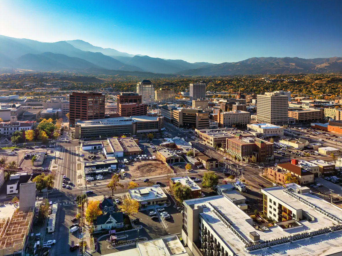 Aerial view of a city with mid-rise buildings, roads, and scattered trees, set against a backdrop of distant mountains under a clear blue sky.