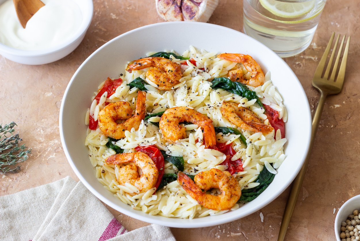 A white bowl filled with orzo pasta, sautéed shrimp, spinach, and tomato pieces, garnished with black pepper. A fork, a glass of white wine, and a bowl of sauce are nearby on a beige surface.