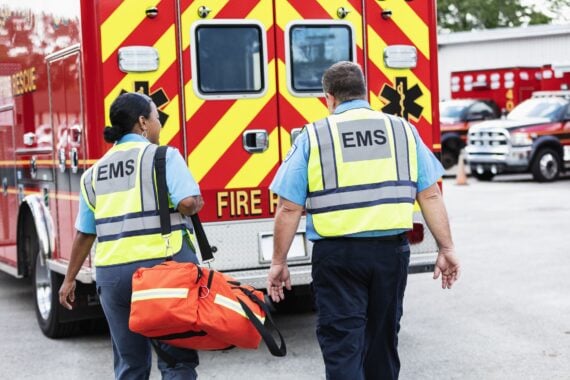 Two EMS workers wearing reflective vests walk toward an ambulance, one carrying an orange medical bag. The back of the ambulance has bold red and yellow stripes. Other emergency vehicles are visible in the background.