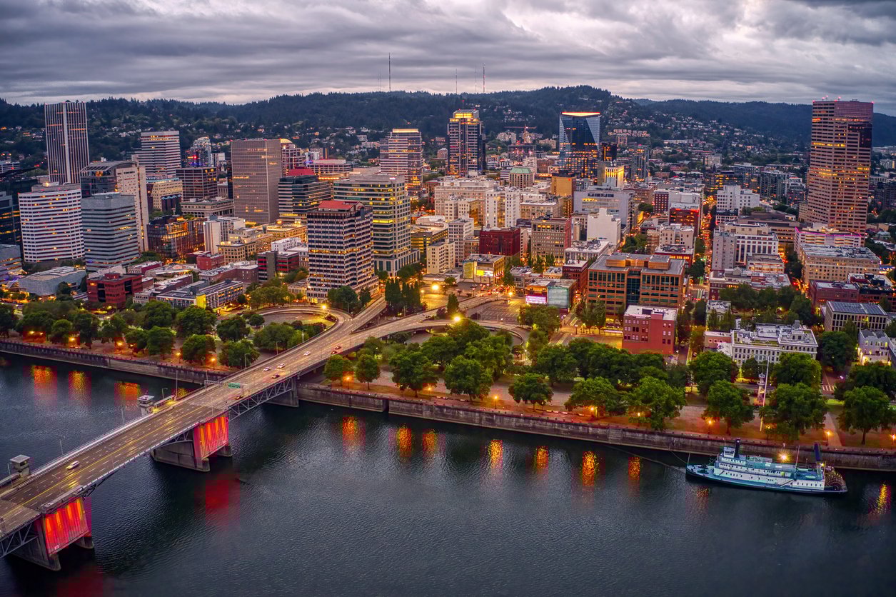 Aerial view of downtown Portland, Oregon at dusk, with illuminated buildings, the Willamette River in the foreground, a lit-up bridge, and tree-lined waterfront under a cloudy sky.