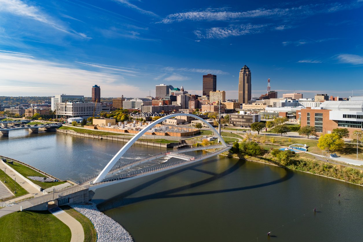 Aerial view of a modern arched pedestrian bridge over a river with a city skyline in the background under a blue sky with scattered clouds.