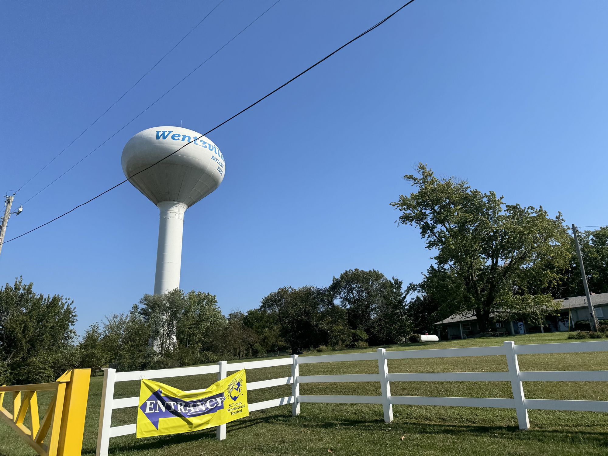 St. Louis, MO, USA - October 14, 2024: A large water tower is visible against a bright blue sky, next to a white fence and greenery, highlighting a quiet suburban area.