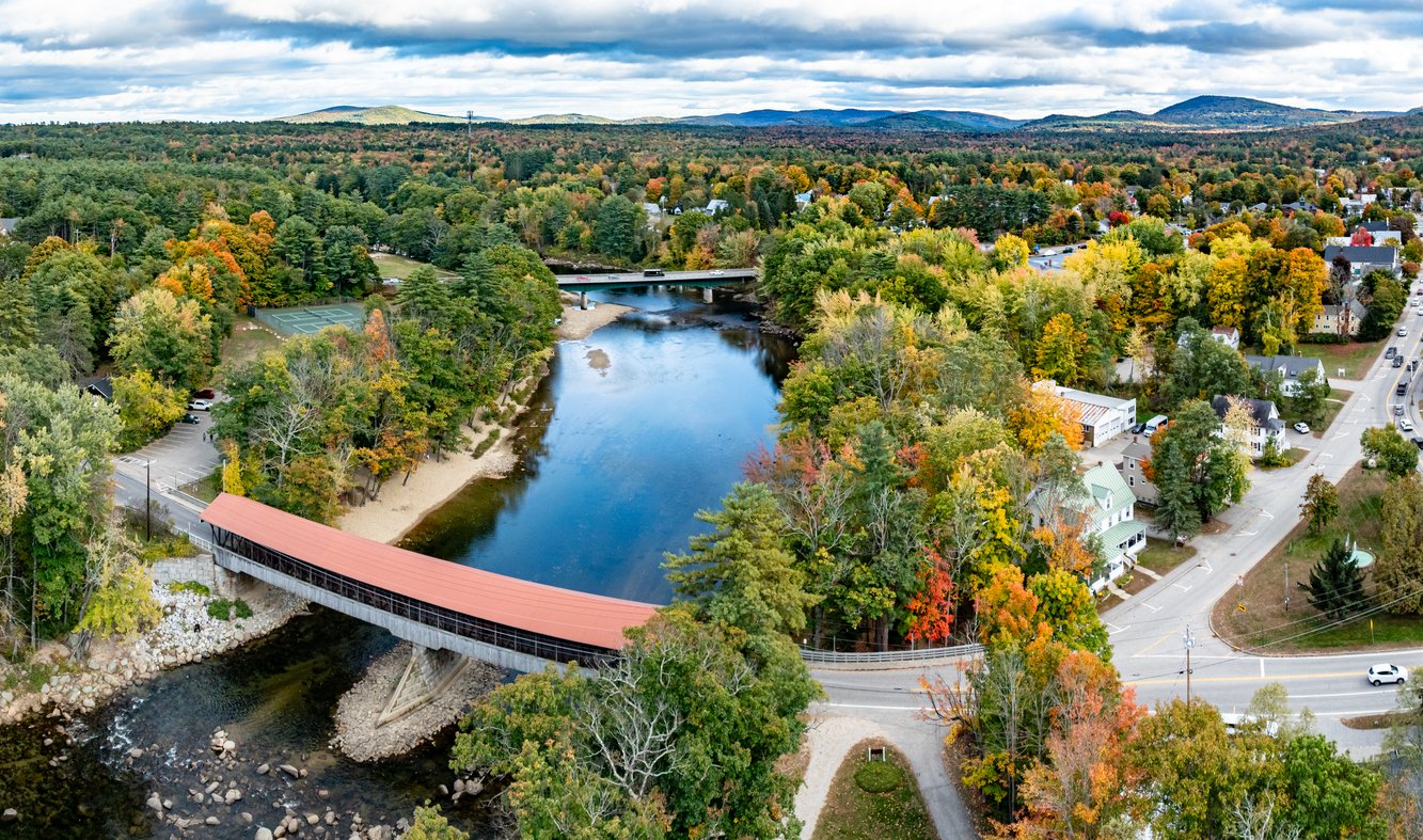 Aerial view of a red-roofed covered bridge crossing a river, surrounded by colorful autumn trees, homes, roads, and distant hills under a cloudy sky.