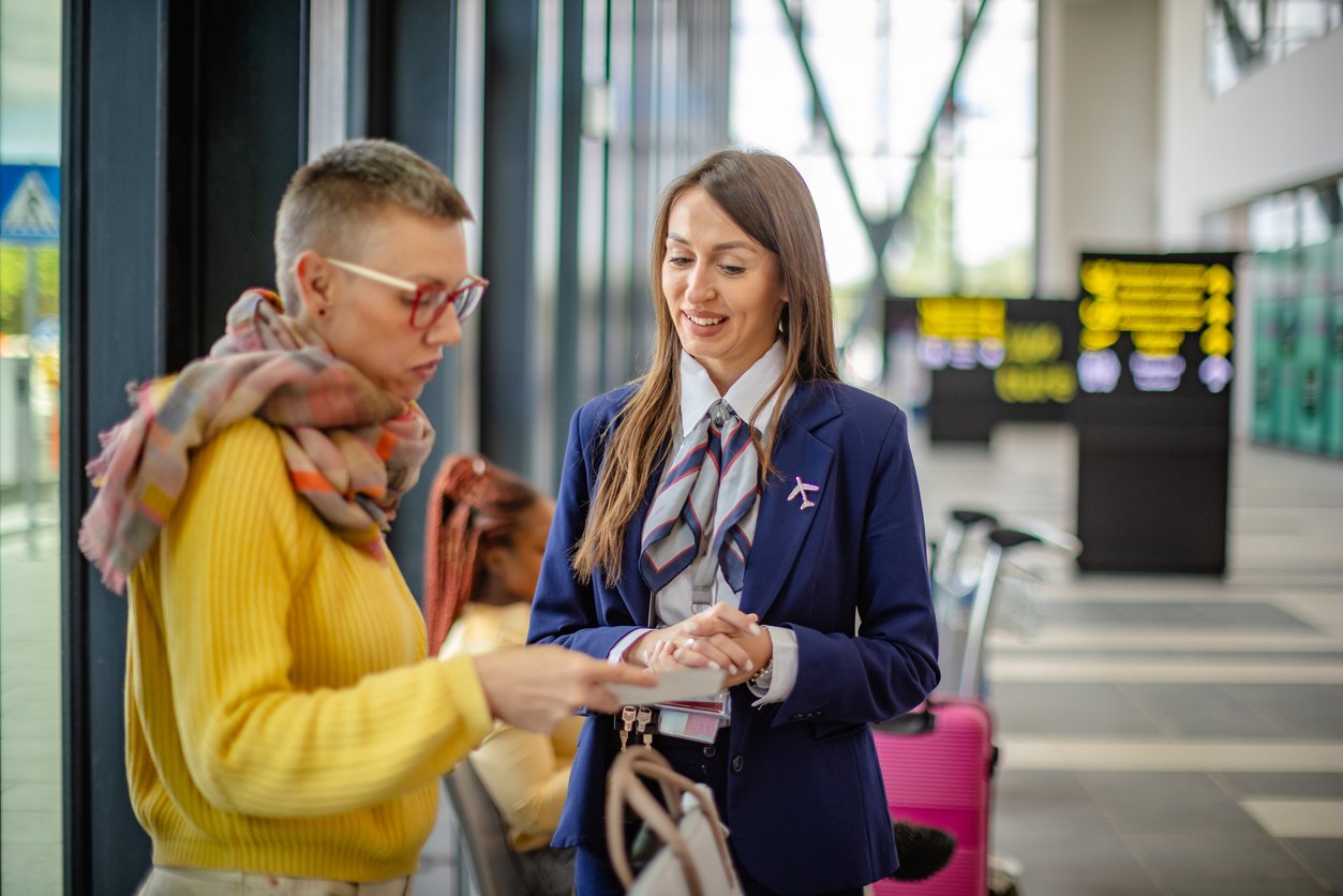 A person in a yellow sweater shows documents to a smiling airline staff member in a navy uniform at an airport, with suitcases and information boards visible in the background.