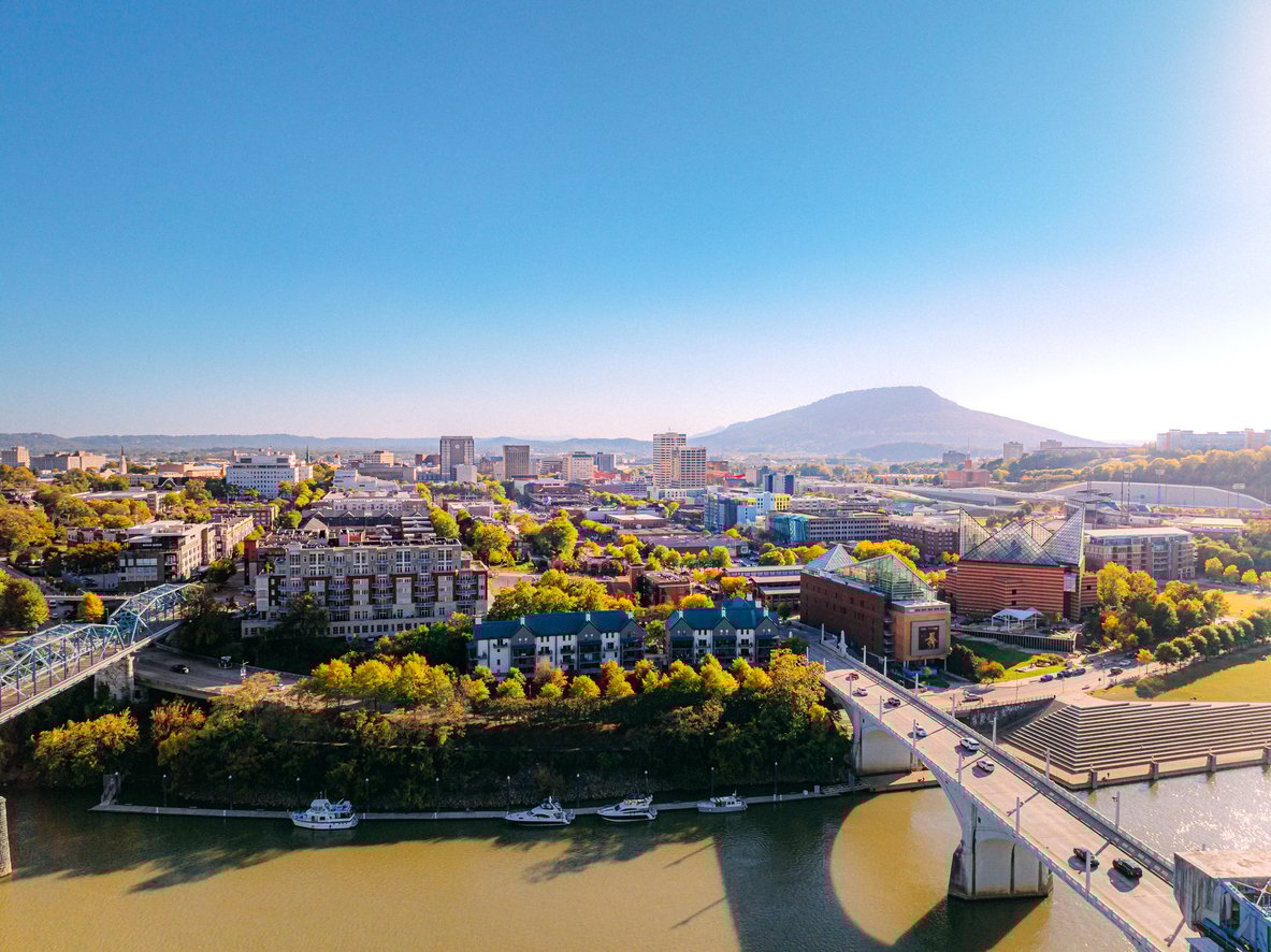 Aerial view of a cityscape with a river, a bridge with cars, colorful buildings, and lush trees. In the background, a large mountain rises under a clear blue sky on a sunny day.