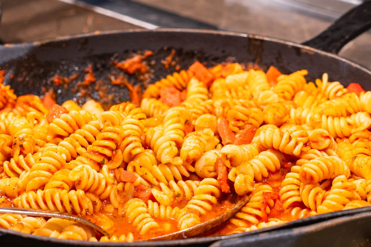 A close-up of rotini pasta covered in a rich tomato sauce with pieces of tomato, served in a black pan with two metal utensils.