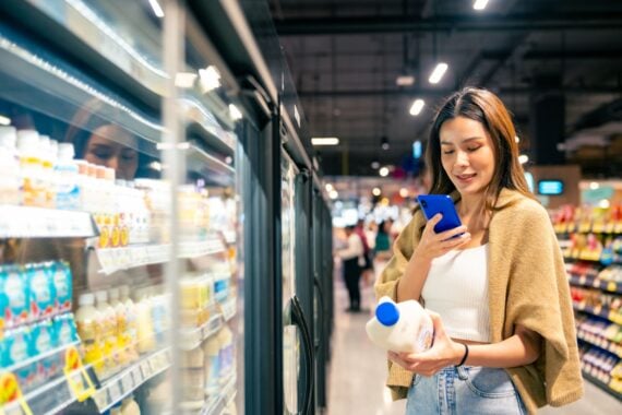 A woman stands in a supermarket aisle, holding a bottle of milk in one hand and looking at her smartphone in the other, while standing next to a refrigerated display of dairy products.