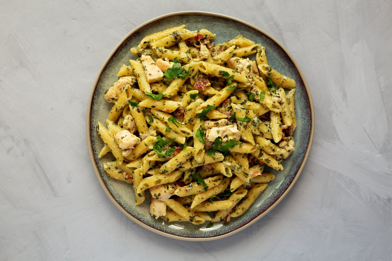 A plate of penne pasta mixed with pieces of chicken, herbs, and garnished with fresh parsley, served on a gray ceramic plate against a light gray background.