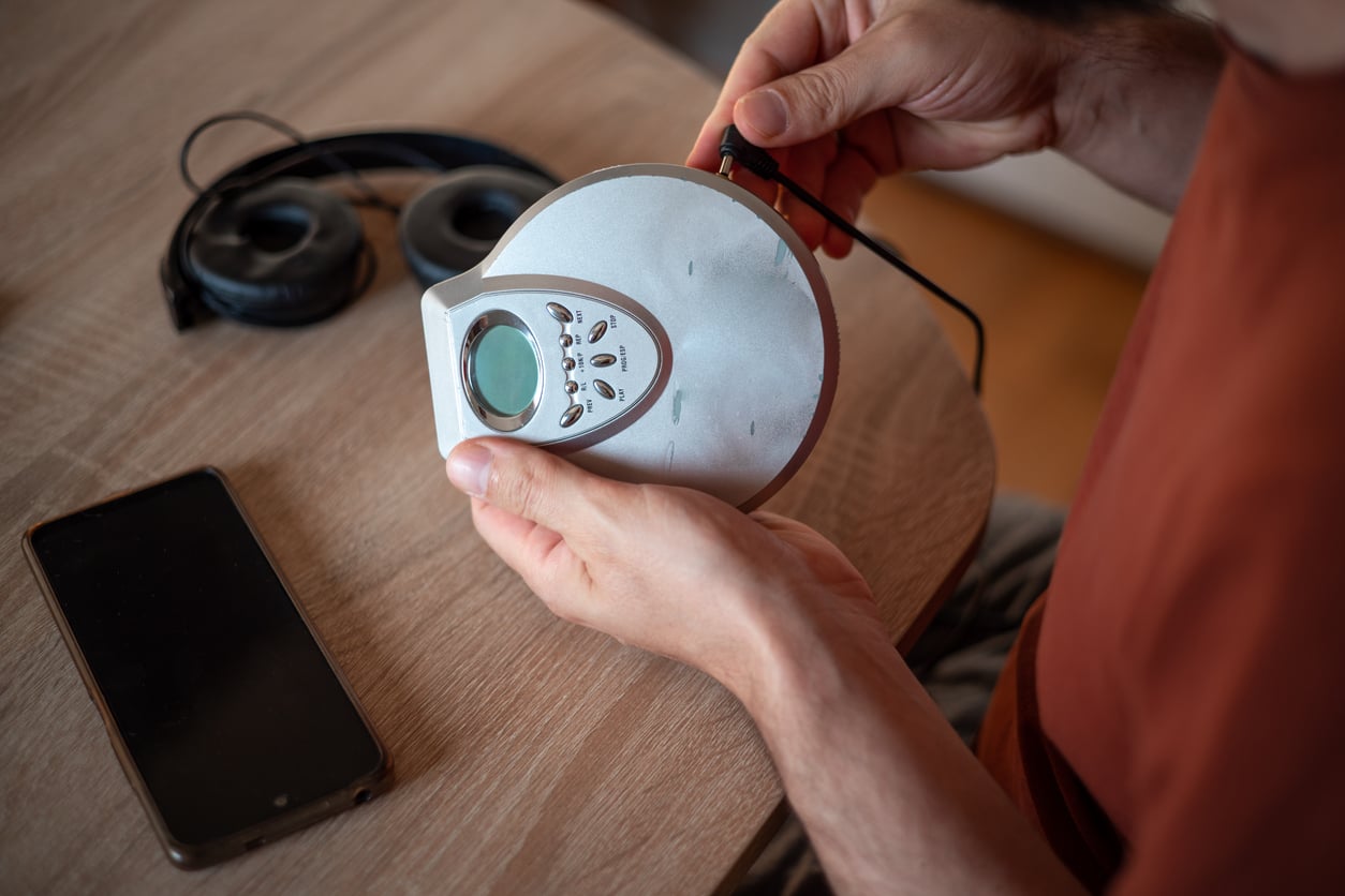 A person holds a portable CD player and plugs in headphones while sitting at a table. A smartphone and a pair of headphones rest on the wooden surface nearby.