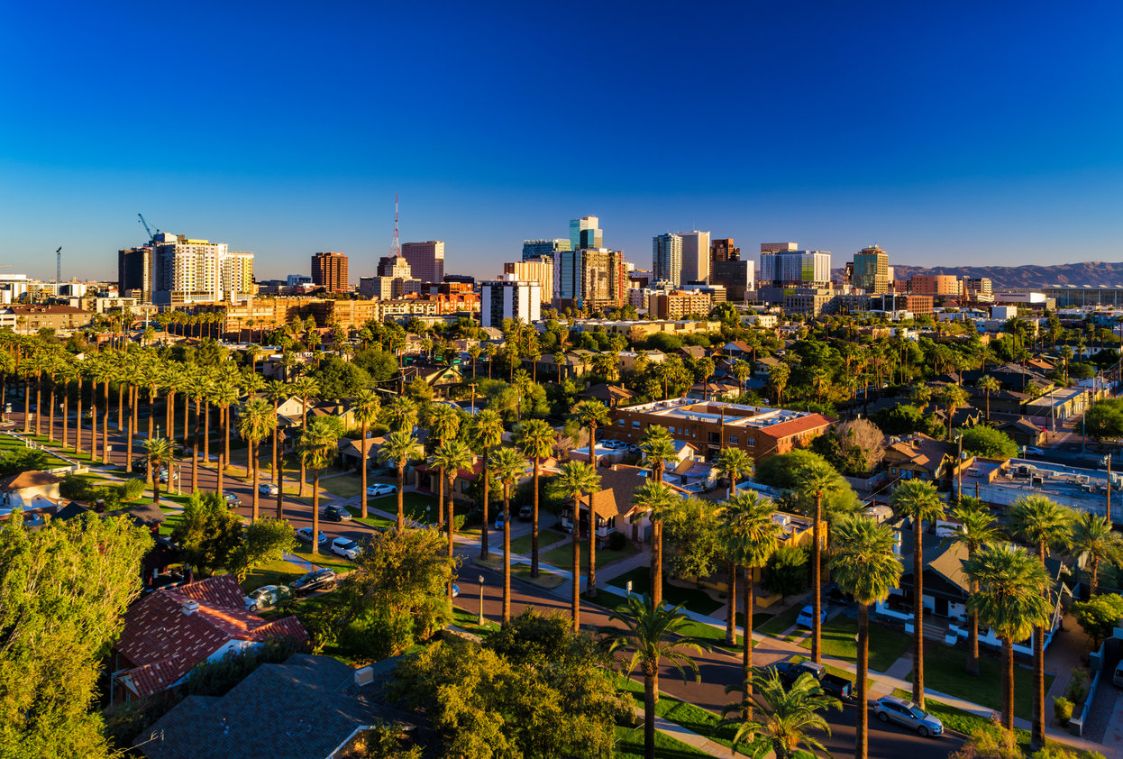 Aerial view of a city skyline with tall buildings and palm trees under a clear blue sky, residential homes in the foreground, and mountains in the distance.