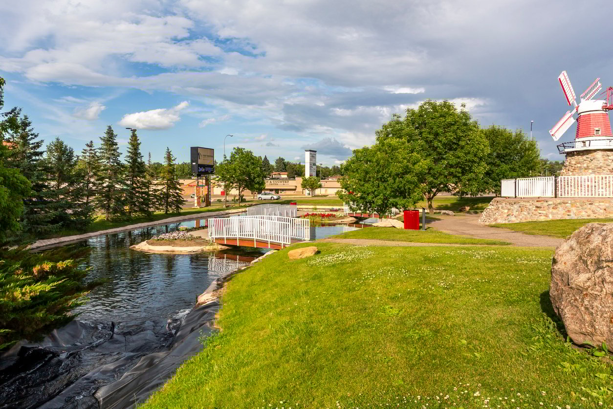 A park with a small canal, a white bridge, green grassy areas, trees, and a red and white windmill under a partly cloudy sky. Buildings and a road are visible in the background.
