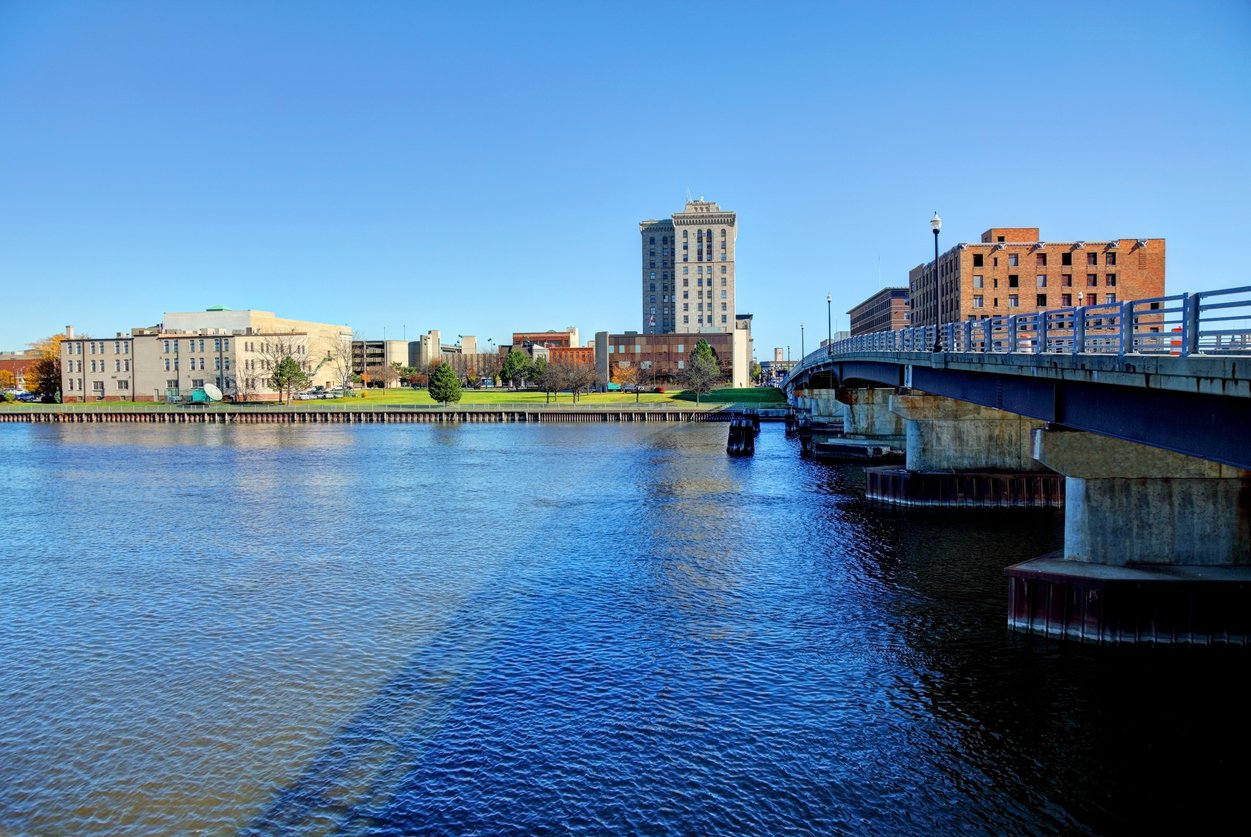 A wide river flows beside a bridge leading into a city with several tall buildings and a clear blue sky overhead.