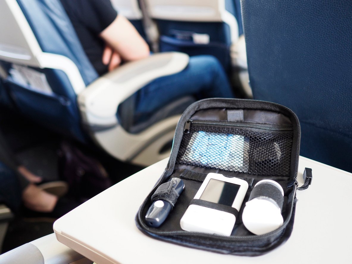 A diabetes kit with a glucometer, test strips, and other supplies sits open on an airplane tray table; a seated passenger is visible in the background.
