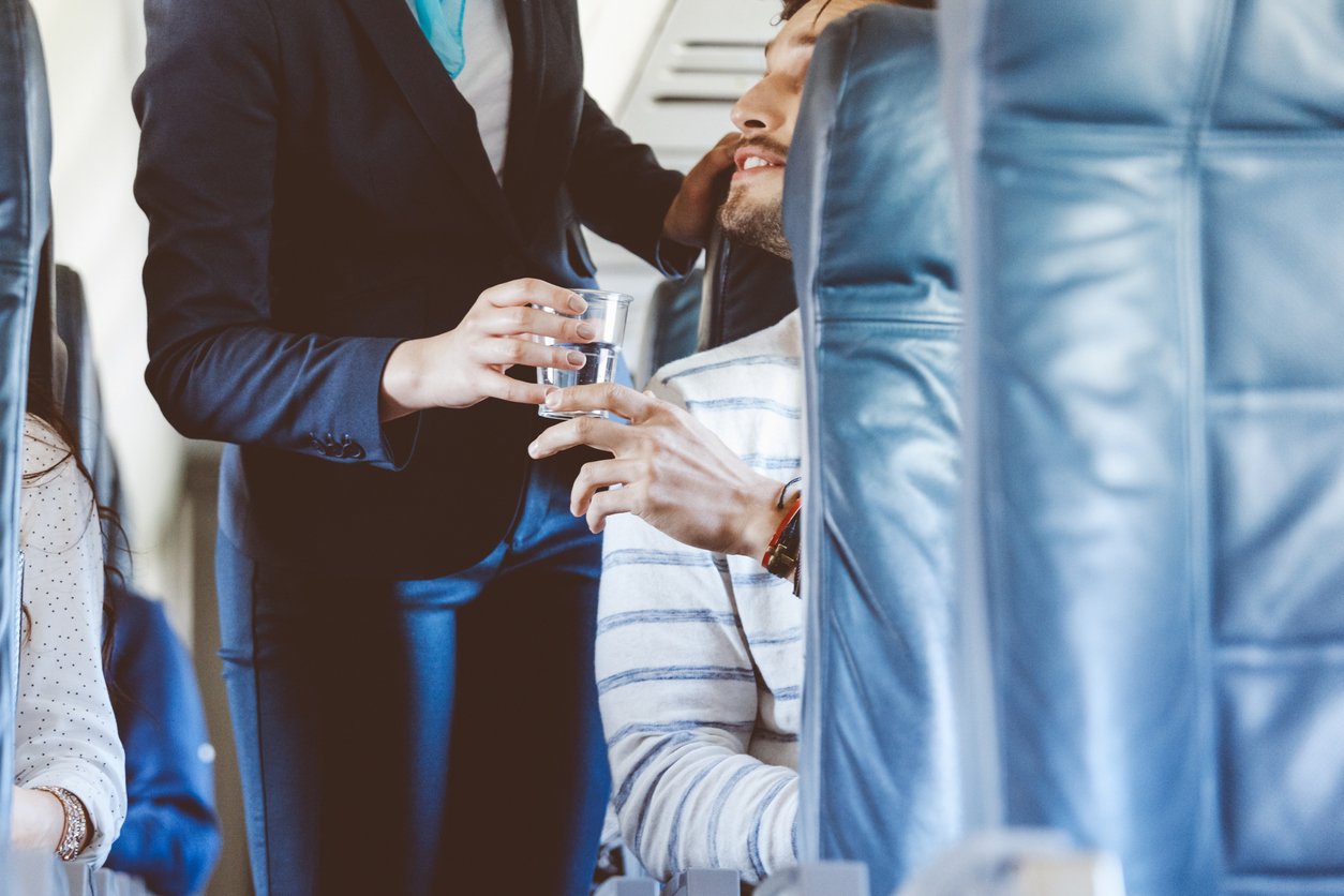 A flight attendant hands a glass of water to a seated passenger on an airplane. The scene is inside the aircraft cabin, with blue seats visible in the foreground and background.