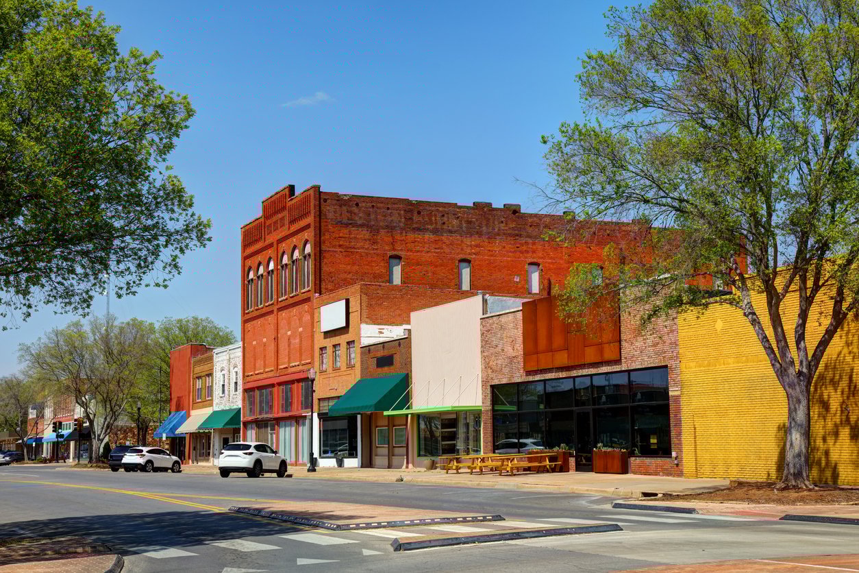 A small-town main street with historic brick buildings, colorful awnings, parked cars, and large leafy trees lining the sidewalk on a sunny day.