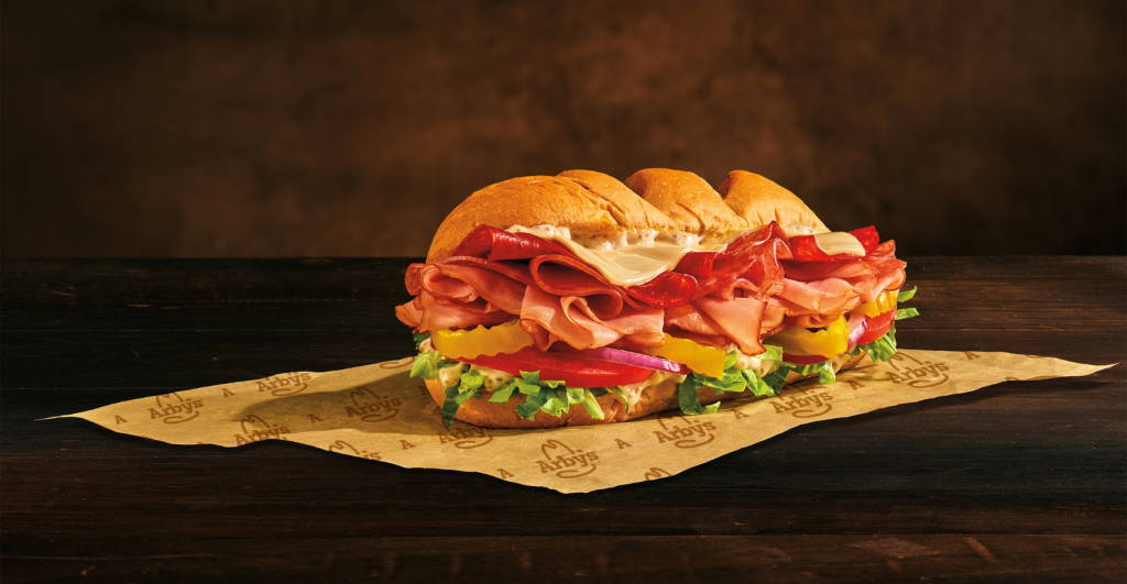 A new fast-food sub sandwich on a wooden surface, piled high with ham, salami, cheese, lettuce, tomatoes, banana peppers, and red onions, all served on an Arby’s branded sandwich wrapper. Loaded Italian