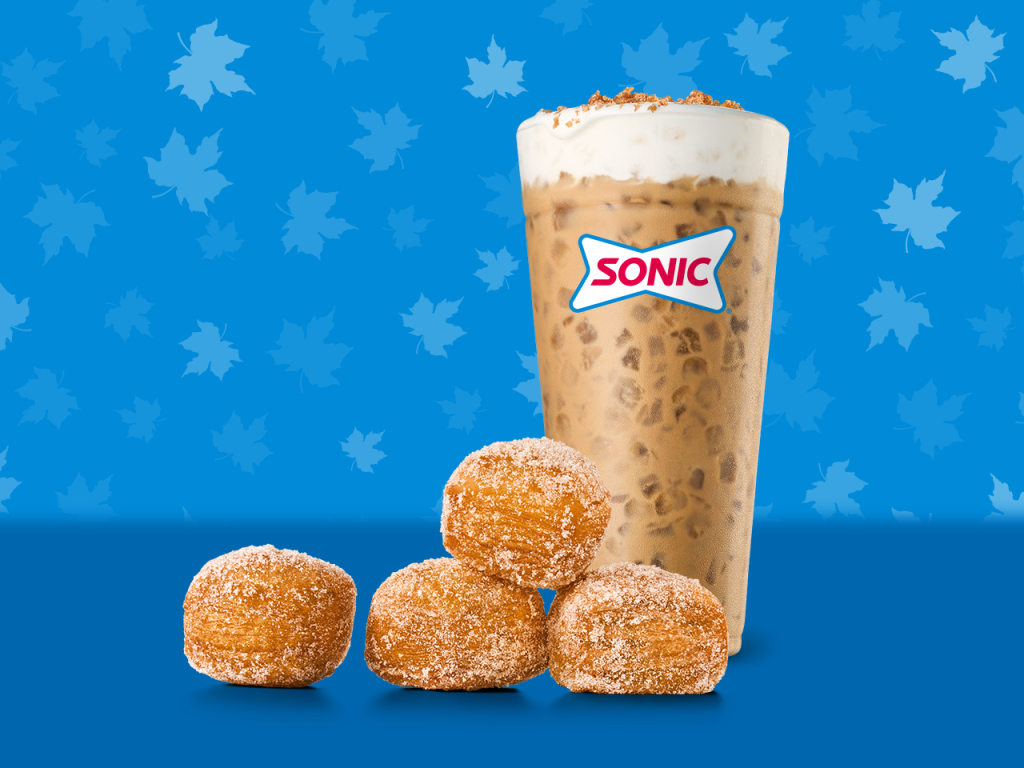 A tall iced coffee with whipped cream and cinnamon on top sits behind five sugar-coated donut holes, all on a blue background with a maple leaf pattern. The cup has a SONIC logo, showcasing this tempting new fast-food treat.