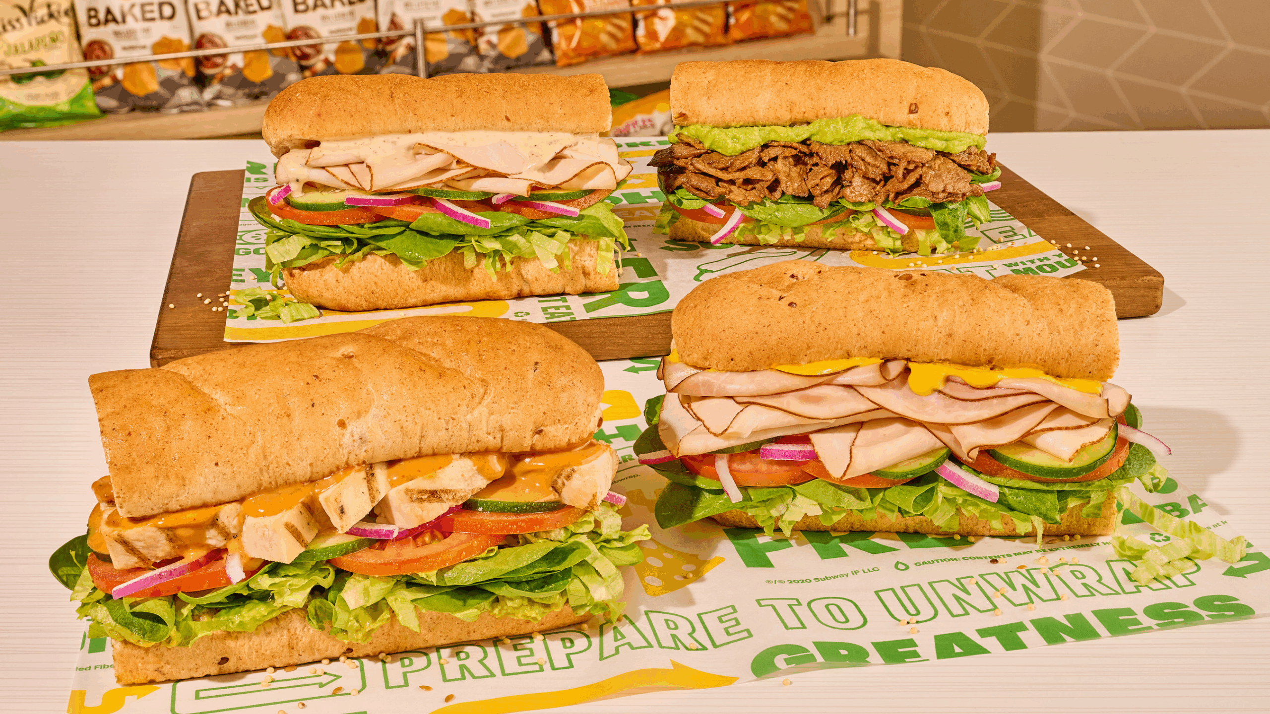 Four sub sandwiches with whole grain bread are displayed on branded sandwich papers. The subs, from a new fast-food spot, feature lettuce, tomato, onions, sliced meats, cheese, and sauces. Bags of chips are visible in the background.