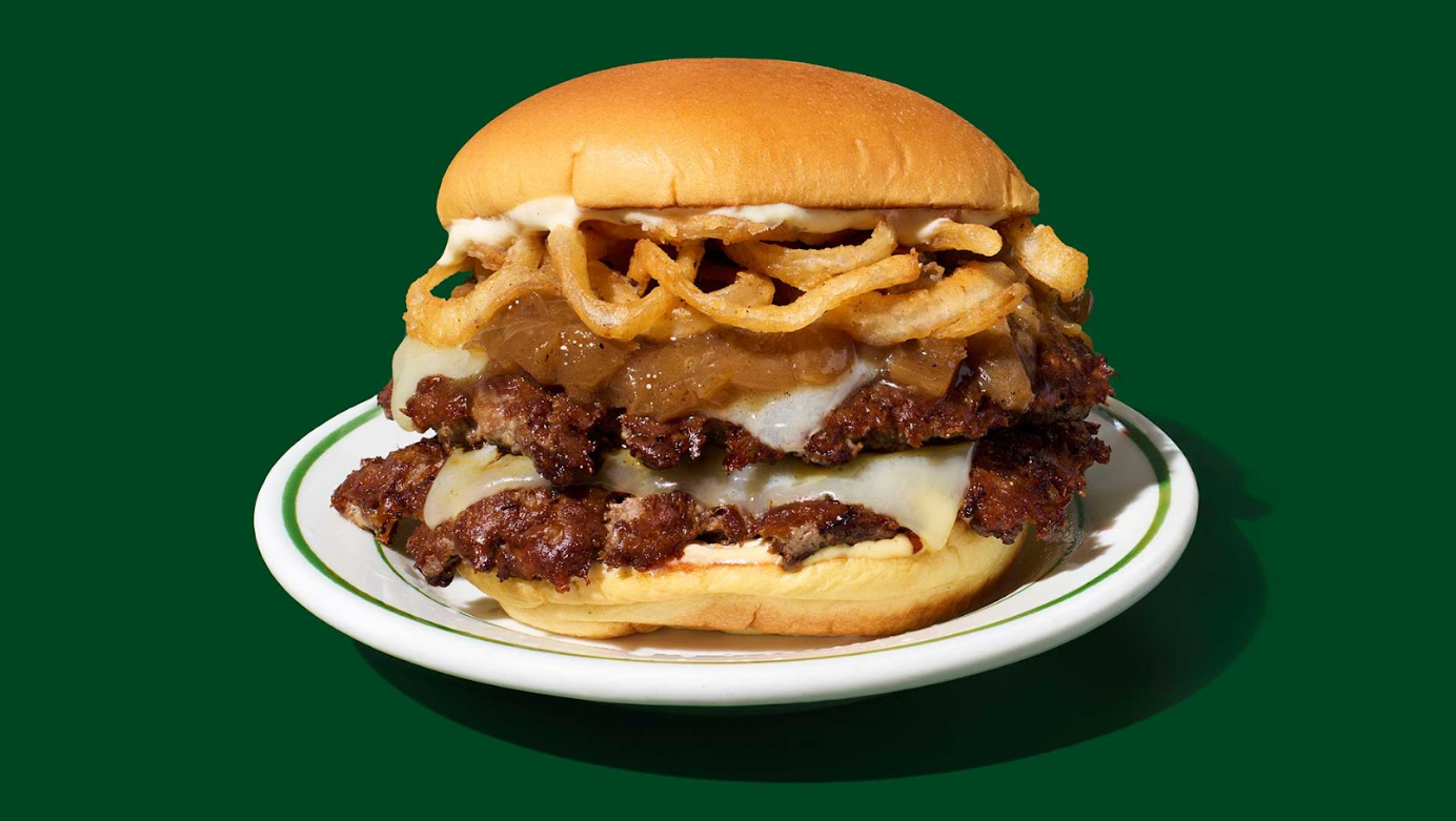 A new fast-food favorite: a cheeseburger with two beef patties, melted cheese, crispy fried onions, caramelized onions, and sauce on a soft bun, served on a white plate against a dark green background. French onion soup burger