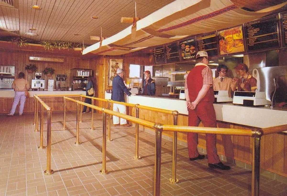 A 1980s fast food restaurant interior with wood paneling, brass railings forming a queue, customers ordering at the counter, and staff in red uniforms. A classic fast-food menu board hangs above the counter.
