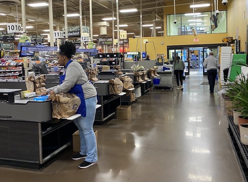 A Kroger grocery store checkout area with a cashier bagging groceries at a register. Several paper bags are on the counters, and a few customers are seen walking in the background. The store appears well-lit and spacious.