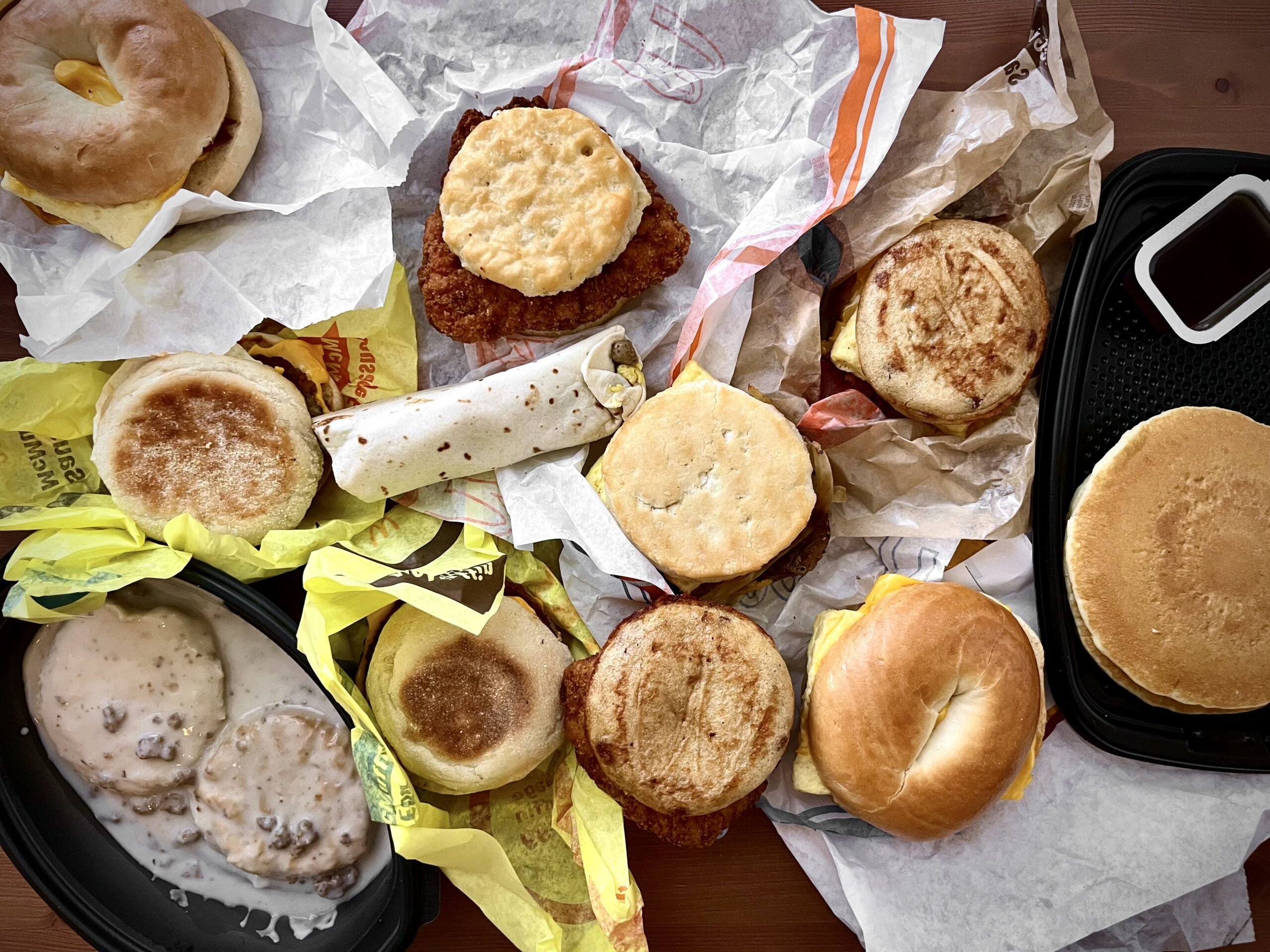 An assortment of McDonald's breakfast items on a table, including biscuits, pancakes, English muffins, a breakfast burrito, gravy with biscuits, and sandwiches wrapped in paper.