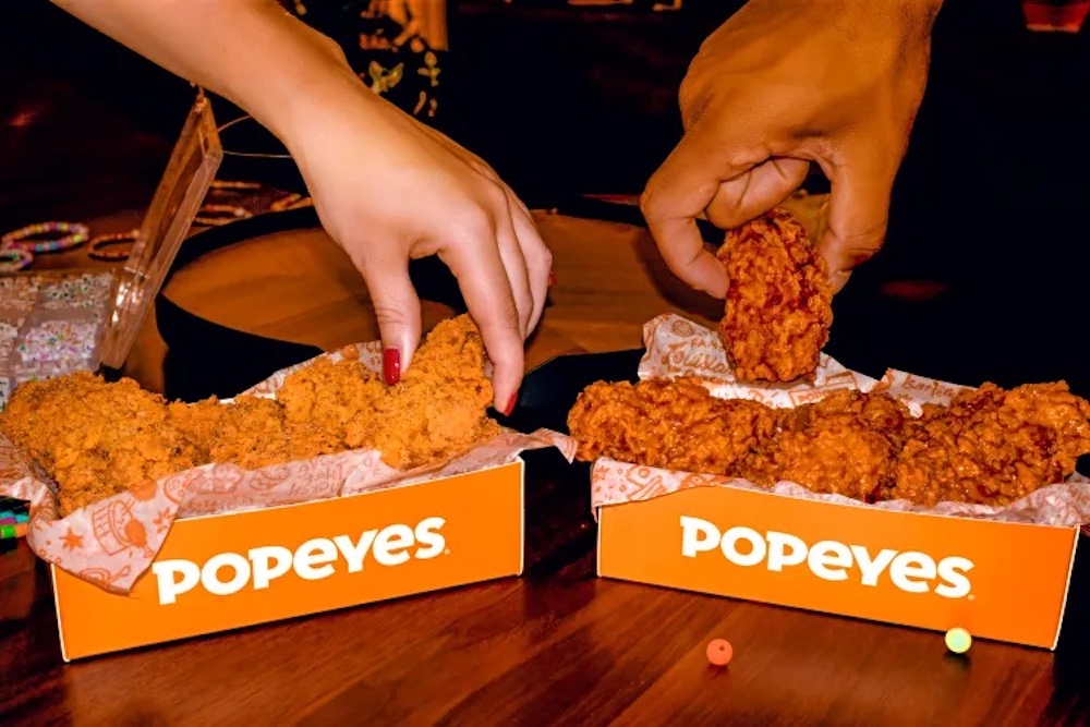 Two people reach for pieces of fried chicken from two orange Popeyes boxes on a wooden table, capturing the joy of sharing a new fast-food meal together.