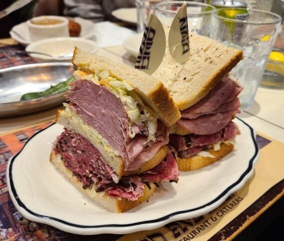 Two thick deli sandwiches with layers of sliced meat and coleslaw on rye bread, each held together by a toothpick, served on a white plate at a restaurant table.