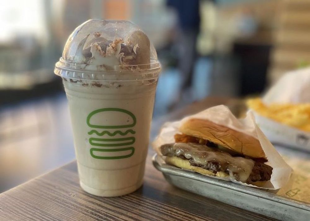 A vanilla milkshake with whipped cream in a clear cup next to a cheeseburger with melted cheese on a metal tray, set on a wooden table in a casual restaurant.