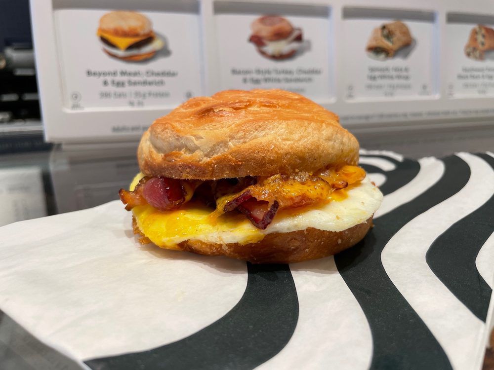 A breakfast sandwich with egg, cheese, and bacon on a croissant sits on a black and white patterned wrapper, with a menu displaying other sandwiches visible in the background.