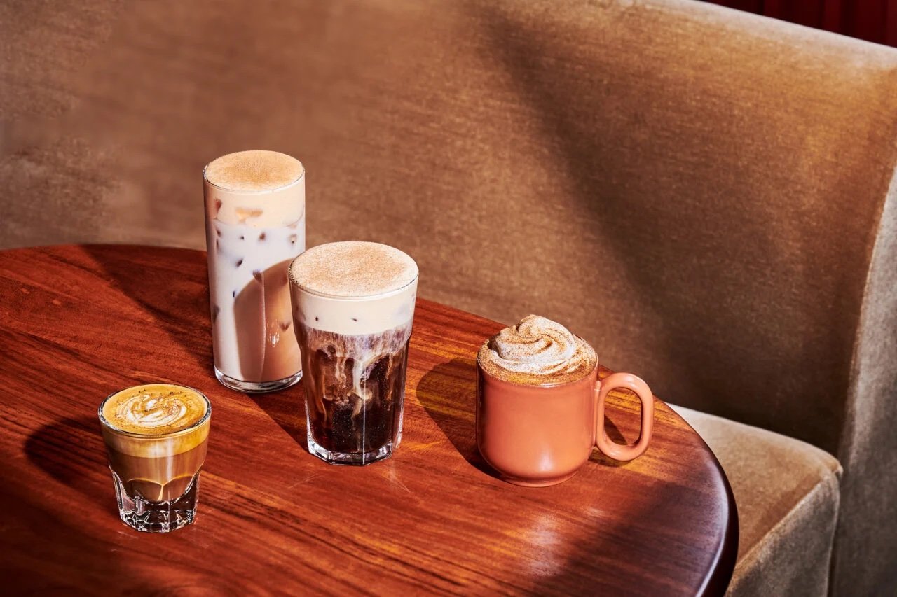 Four coffee drinks, including a small espresso, two iced coffees in tall glasses, and a cappuccino in a copper mug, are arranged on a round wooden table—bringing new fast-food style energy to the cozy tan upholstered bench in the background.
