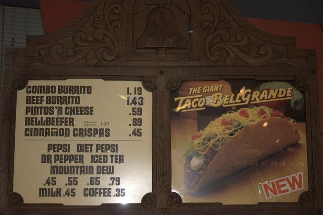 A vintage Taco Bell menu with wooden framing displays food items and prices on the left, and an advertisement for "The Giant Taco BellGrande" with a taco image on the right.