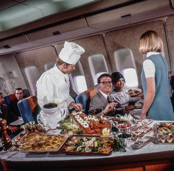 Passengers on an airplane are served gourmet food by a chef and a flight attendant. Platters of elaborate dishes cover the cart, and people look happy and engaged in conversation.