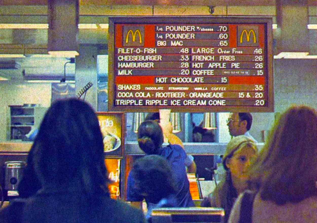 Customers stand in line at a McDonald’s counter, looking up at a vintage fast-food menu board featuring classic items and old prices like 70-cent Quarter Pounders and 48-cent Filet-O-Fish.