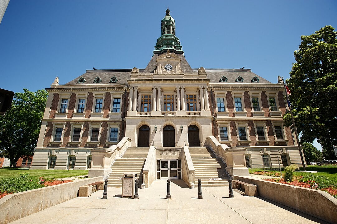 A large, historic courthouse building with a clock tower, columns, and grand stone steps leading to the entrance, set against a clear blue sky and surrounded by green trees and manicured lawns. Hall County Courthouse in Grand Island, June 2014

