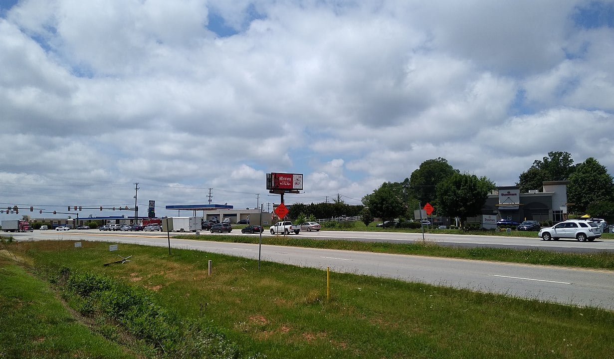 A wide roadside view shows a busy intersection with cars stopped at a traffic light, businesses, and storefronts in the background under a partly cloudy sky. A grassy area and two orange construction signs are in the foreground.